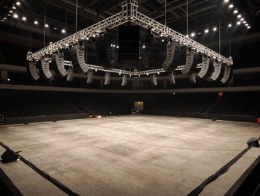 Wide panoramic shot of a large arena floor during setup, rows of line-array speakers suspended from a central truss, the floor empty and lit only by the work lights, cable runs visible along the stage lip, the venue's upper deck in dim silhouette