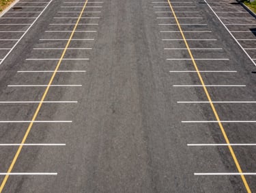 High-angle drone shot of a massive, perfectly striped empty commercial parking lot with crisp white and yellow lines, stark midday sun.