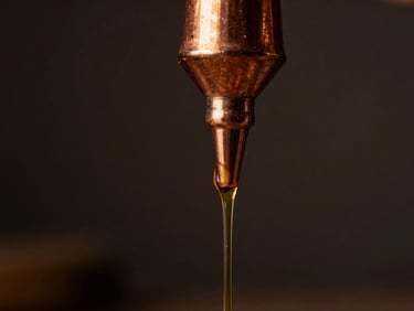 Macro close-up of thick golden amber oud oil slowly dripping from a traditional copper still nozzle, dark moody background, low-key lighting.