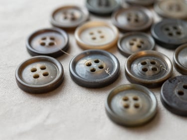 Macro close-up of vintage buttons and worn-in threads on a light neutral linen surface, soft diffused morning lighting.