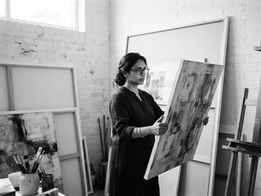 High-contrast black and white medium format portrait of an Indian fine artist in a sunlit studio, surrounded by large stretched canvases, soft natural light from a high window, minimalist composition.