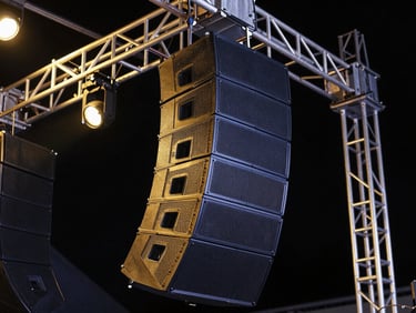 Close-up of professional line array speakers hanging from an aluminum truss, dramatic stage lighting in the background, 35mm