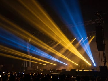 Dramatic concert lighting show with gold and blue laser beams cutting through haze over a stage, wide environmental shot