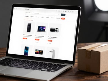 Tight close-up of a laptop screen showing an e-commerce product category page with structured product grid and breadcrumb navigation, placed on a dark wooden surface with a small cardboard shipping box beside it, clean directional studio light from the upper right