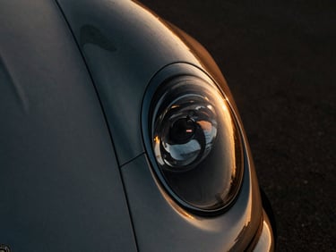 Extreme close-up of a vintage luxury sports car headlight and sculpted fender, dramatic golden hour reflections, dark moody atmosphere