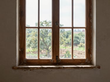 Wide interior shot of a restored original wooden window frame, natural daylight streaming through restored glass panes onto worn plaster walls, construction dust settled on sill, honest mid-restoration detail, no staging