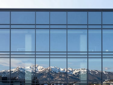 Wide shot of a minimalist corporate glass facade reflecting a bright blue sky and distant snow-capped mountain peaks, clean lines, high contrast.