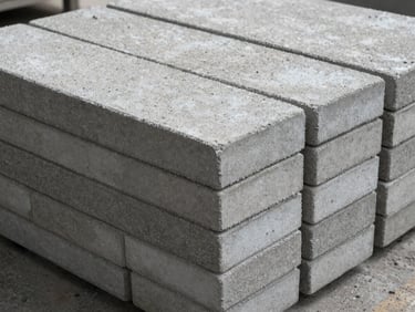 Close-up of concrete paving stones stacked in two courses on a factory floor, textured top surface with fine aggregate, natural overcast light from above, side profile shows uniform thickness