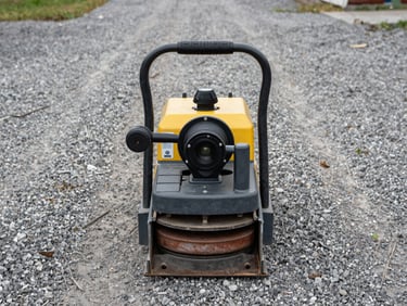 Operator-angle shot of a plate compactor on a gravel surface, viewed from behind-above, compactor plate and handle visible, diffused daylight from an open yard, no people