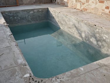 Wide shot of an existing pool mid-renovation in Alicante, old coping removed and structural shell exposed, natural afternoon light revealing concrete surface and drainage channels, cool documentary tone