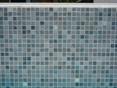 Close-up of freshly applied gresite mosaic tile on a pool wall, cool grey-blue tones, water line visible at the top edge, natural midday light, documentary framing showing material texture and craft detail