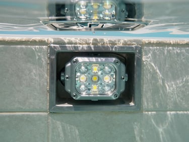 Close-up of a pool technical installation — underwater LED light fitting recessed into tiled wall, stainless steel drain cover, clear water above, sharp midday Mediterranean light, technical documentary framing