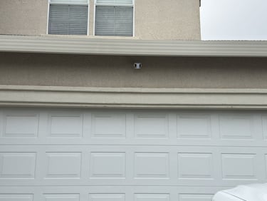 A white sectional garage door under a two-story tan stucco house with a modern security camera installed.