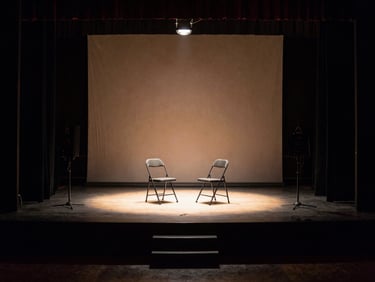 Small bare stage with two folding chairs and a single work light, viewed from the wings, deep shadows framing the lit performance area