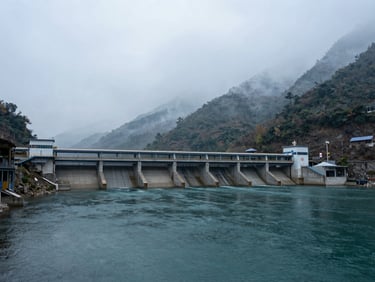 Wide environmental landscape shot of a concrete run-of-river hydroelectric plant integrated into a misty mountain river basin, overcast daylight