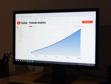 Desktop monitor showing a YouTube analytics dashboard with a subscriber and view count growth chart spiking upward over four months, screen-glow lighting, surrounding desk in near-darkness