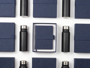 Clean overhead flat-lay of premium linen-wrapped gift boxes, open to reveal debossed journals and matte black thermal flasks, soft studio lighting