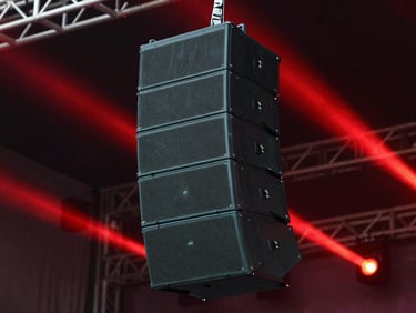 Close-up of high-end line array speakers hanging from a truss, dramatic outdoor concert stage background with crimson light beams, sharp focus