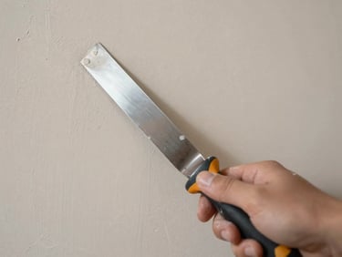Overhead close-up of fresh plaster being applied to a drywall panel, wide joint knife in frame, natural studio daylight, neutral warm tones, no people visible