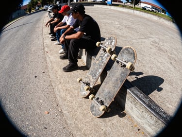 A group of skaters sitting on a concrete curb next to a dusty road, worn skateboards leaning against them, harsh street lighting.