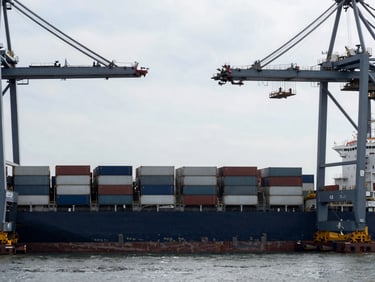 Wide shot of a container ship docked at a port terminal, crane structures framing the sky, steel grey tones.