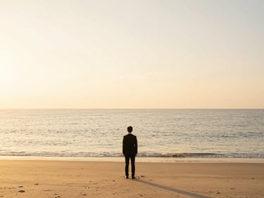 A solitary figure in professional attire standing on a pristine beach at dawn, looking out at the vast ocean horizon, soft golden light, expansive negative space.