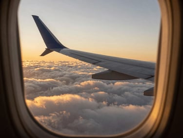 An airplane wing cutting through soft golden clouds during sunrise, captured from a premium cabin window.