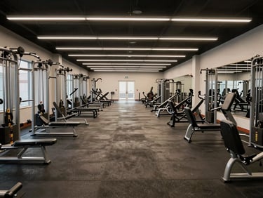 Wide-angle shot of an unoccupied gym floor, exercise equipment in neat rows, overhead lighting neutral and even, full room depth from entrance to back wall, no people