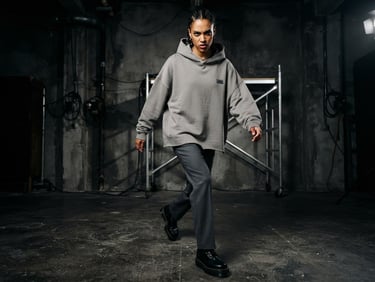 Low-angle dynamic studio shot of a model wearing an oversized concrete-gray streetwear hoodie and tailored trousers, high-contrast industrial lighting, dramatic shadows, sharp focus, 35mm lens