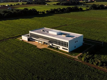 Dramatic high-contrast aerial shot of a modern farm headquarters in Bahia, surrounded by lush green cultivated fields under high afternoon sun