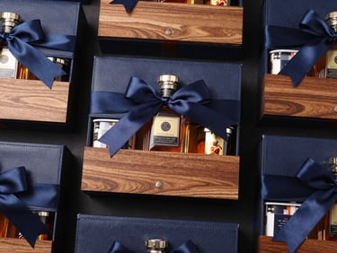 Overhead flat-lay of a luxury corporate gift hamper with high-texture materials like leather, wood, and silk ribbons, featuring a premium spirit bottle and gourmet items.