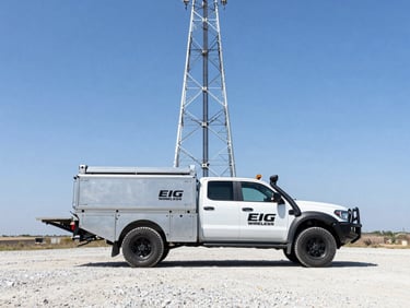 Wide environmental shot of a branded EIG Wireless fleet vehicle, a heavy-duty pickup truck with a utility bed, parked on a gravel access road at the base of a tall cell tower. Bright, high-contrast daylight, industrial editorial photography, steel and sky tones.