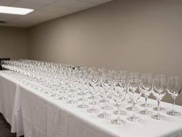 Wide shot of a corporate hospitality drinks reception setup, rows of filled wine and water glasses on a white-draped table against a neutral wall, clean overhead lighting, no staff or guests