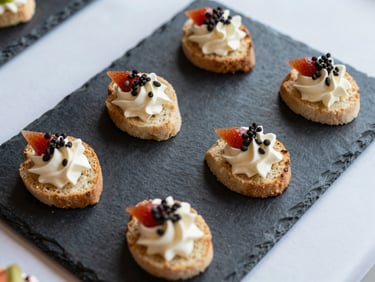 Overhead close-up of a private event canapé tray, six uniform bite-sized items on a slate board, clean daylight, white preparation surface visible at edges