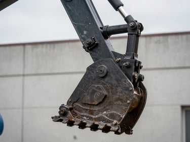 Close detail shot of heavy machinery hydraulic arm and bucket teeth, overcast diffused daylight revealing surface wear and grease fittings, frame tight on the mechanical joint showing scale and working condition, dark steel tones against a pale concrete background