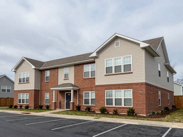 Wide exterior shot of a two-story residential apartment building, natural overcast light, brick and siding facade, parking area visible in foreground, no people, factual documentary framing