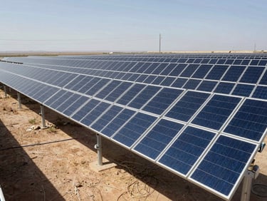 Wide environmental shot of a utility-scale solar field, rows of photovoltaic panels stretching to the horizon, overhead power lines visible in background, harsh midday sun casting short shadows, desert terrain