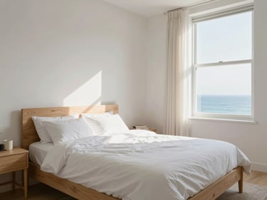 Bright, airy bedroom in a coliving suite. Natural light from a large window illuminates soft white bedding and light wood furniture. Minimal decor. A coastal view visible through the window. Shot from corner to emphasize spaciousness.