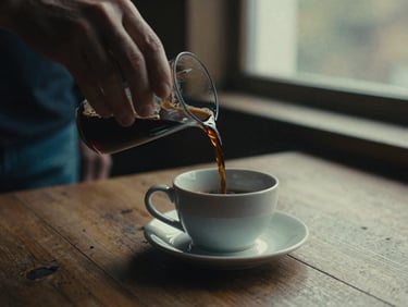 Cinematic close-up of hands pouring dark rich coffee into a ceramic cup on a rustic wooden table, soft dramatic window lighting, warm moody tones, 35mm grain