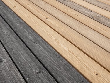 Timber deck close-up showing half-cleaned boards, dark weathered grey on one side giving way to clean natural wood grain on the other, natural overcast light