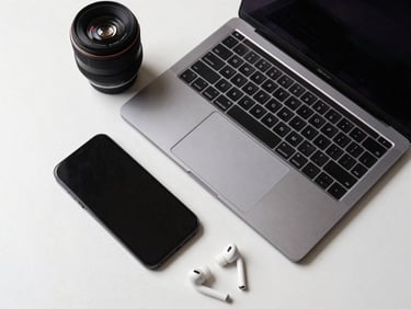 Overhead flat-lay of a modern laptop open beside a smartphone and wireless earbuds on a clean white surface, neutral studio lighting, sharp spec detail