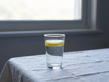 A glass of water with a lemon slice on a white linen tablecloth, soft morning window light, serene minimalist setting