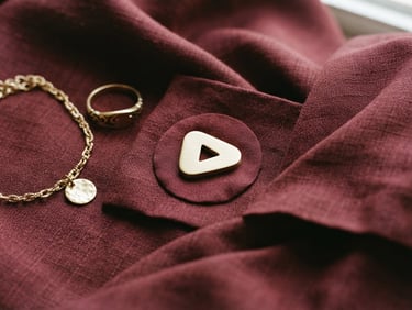 A close-up of a YouTube play button icon, subtly integrated into an elegant flat lay of silk fabric and a gold accessory, soft natural window light, rich burgundy and gold tones, 35mm