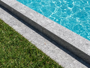 Neatly edged green garden borders meeting grey granite pool coping stones, crisp morning light showing dew, sharp architectural lines.