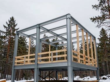 Exposed structural steel frame of a modern cabin under construction in a snowy pine forest, low-angle shot showing massive steel connections, overcast sky.