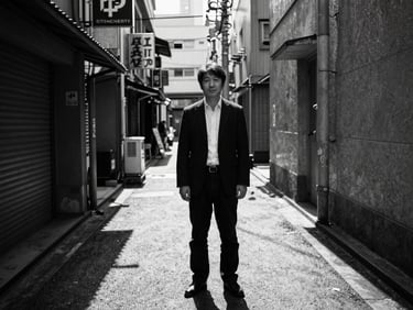High-contrast black-and-white wide-angle portrait of a male curator standing in a Tokyo alleyway, deep shadows, direct sunlight, raw urban texture.