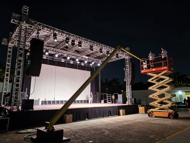Wide shot of a stage rig being assembled inside a Miami venue at night, crew on scissor lifts adjusting beam lights, cables and truss in foreground, dramatic contrast between lit stage and dark house, high-contrast night photography