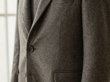 Close-up of a textured warm gray wool blazer, showcasing the pocket detail and natural fabric texture under soft window light