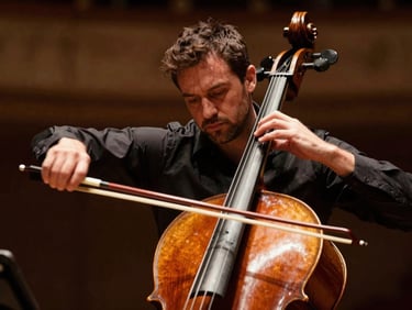 Close frame of Paolo Tedesco's bow arm mid-stroke on cello strings under warm amber stage lighting, motion visible in the bow hair, dark concert hall background