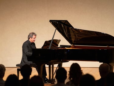 Stage-level view of Paolo Tedesco performing with piano accompanist, both figures lit by warm proscenium lighting, audience silhouettes in foreground
